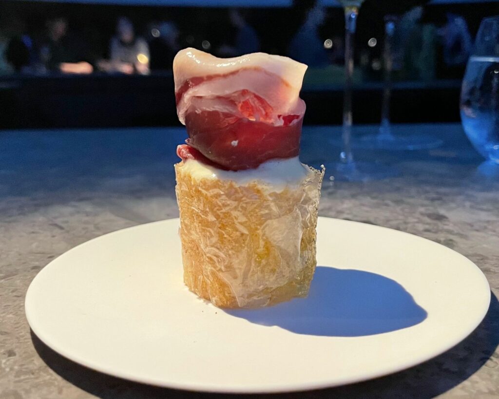 Airy bread dell'Alchemist: un pane leggerissimo fatto con fogli di fecola di patate spennellati con burro dorato, lievito tostato e succo di cipolla. Viene arrotolato come un croissant e cotto sottovuoto per creare una consistenza gonfia e croccante. È guarnito con una schiuma a base di lievito, tuorli d'uovo al burro, crème fraîche e sottili fette di prosciutto Joselito Pata Negra annata 2017.