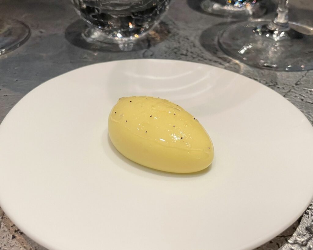 Perfect omelette dell'Alchemist: una membrana fragile di tuorlo modellata in uno stampo 3D, riempita con crema di tuorlo d'uovo e formaggio Comté, guarnita con una fettina di Joselito di 0,5 mm di spessore, decorazione con un motivo al tartufo nero e spennellata di burro aromatizzato al pepe nero tramite ultrasuoni
